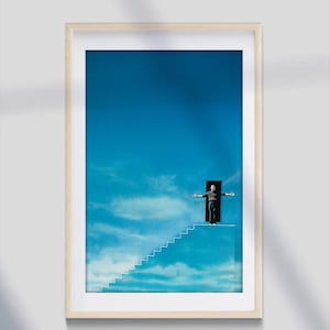 May include: A framed print depicting a man standing on a staircase leading up to a doorway in the sky. The sky is blue with white clouds. The man is wearing a dark shirt and trousers. The staircase is white and the doorway is black.