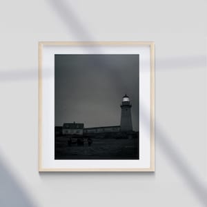 May include: Framed monochrome photograph featuring a lighthouse with a lit beacon, a small building, and figures on the ground. The sky is overcast. The frame is light-coloured wood.