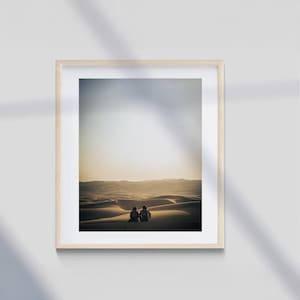 May include: Framed photograph of a desert landscape with two figures seated on a sand dune. The image is bathed in warm sunlight, with soft shadows and a light wooden frame.