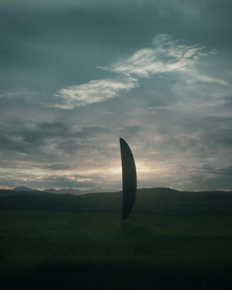 May include: A dark, curved, vertical object hovers over a green field, with hills and a cloudy sky in the background. The object's form resembles a blade or a crescent, with a dark, solid hue. The scene creates a sense of mystery and intrigue.