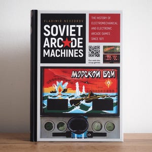Può includere: Un libro intitolato "Soviet Arcade Machines" di Vladimir Nevzorov. La copertina presenta una stella rossa e il testo "Soviet Arcade Machines" in lettere bianche su sfondo nero. Il libro presenta anche una grafica di gioco arcade colorata.