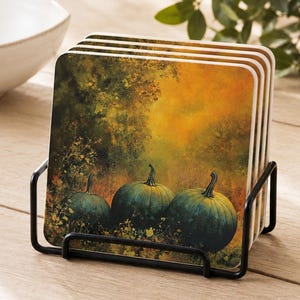 May include: A set of four square coasters held in a black metal wire stand. Each coaster features a printed image of three dark green pumpkins resting in a field against a backdrop of warm, golden-toned autumn foliage and trees.