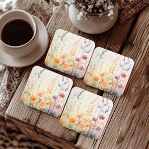 May include: Four square coasters with a watercolour floral design featuring orange, purple, and yellow flowers. The coasters are arranged on a rustic wooden surface, with a cup of coffee and a small vase.