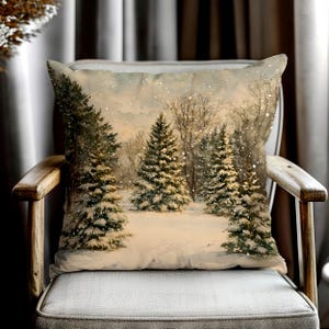 May include: A square throw pillow featuring a printed watercolor illustration of a snowy winter forest scene. The design depicts several evergreen pine trees covered in white snow, set against a pale, overcast sky with falling snowflakes.
