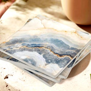 May include: A stack of four square coasters with a marbled design. The coasters feature a blend of white, blue, and gold tones, creating a natural, stone-like appearance. The coasters are stacked on top of each other.