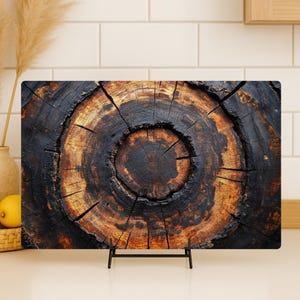 May include: Rectangular cutting board featuring a close-up of a tree trunk cross-section. The wood grain shows concentric rings in shades of brown and black, with visible cracks. The board is displayed on a black metal stand, suitable for kitchen use.