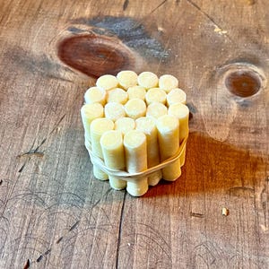 May include: A bundle of small, cylindrical, light yellow objects, held together by a tan rubber band. The objects are arranged in a compact, rectangular shape. The background is a wooden surface with visible grain.