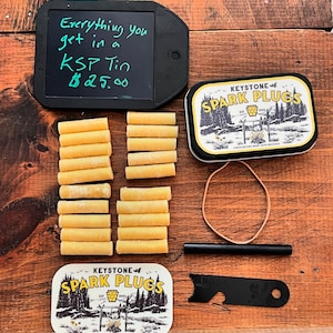 Può includere: Una composizione di oggetti su una superficie di legno, tra cui un tablet nero con testo scritto a mano, due scatole con l'etichetta "Keystone Spark Plugs" e diversi oggetti cilindrici gialli. Sono inclusi anche un'asta nera, un multiutensile nero e un elastico. Il tablet recita "Everything you get in a KSP Tin $25.00".