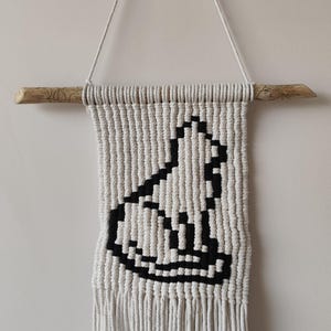 Peut inclure: Suspension murale en macramé avec un motif géométrique noir et blanc. L'œuvre est suspendue à une cheville en bois naturel et comprend une frange en bas. Le motif est une image pixélisée d'un chat.