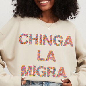Könnte beinhalten: Ein beiger Pullover mit dem Spruch "CHINGA LA MIGRA", der in bunten Blumen geschrieben ist.