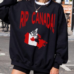 Puede incluir: Sudadera negra con un gráfico de bandera canadiense roja y blanca y el texto "RIP CANADA!" impreso en la parte delantera.