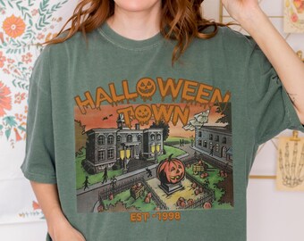 May include: Green t-shirt featuring a Halloween Town graphic with a pumpkin and buildings. The design includes the text "HALLOWEEN TOWN" and "EST - 1998". The shirt is a relaxed fit.