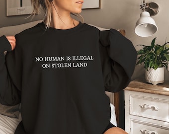 Könnte beinhalten: Schwarzer Pullover mit dem Text "No Human is Illegal on Stolen Land" in Weiß.