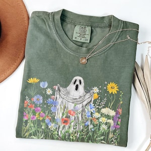 May include: Olive green t-shirt with a ghost graphic in a field of colorful flowers. The shirt has the Comfort Colors brand label and a small gold necklace. The shirt is a size small.