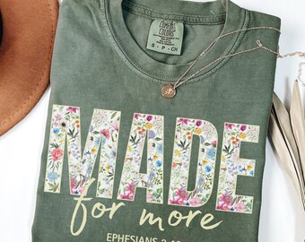 May include: Olive green t-shirt with the words "MADE for more" in a floral design. The text "Ephesians 2:10" is printed below. A gold necklace with a pendant is draped on the shirt.