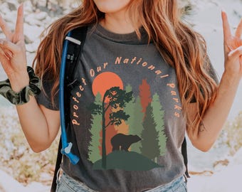 May include: A dark grey t-shirt with a graphic print of a bear in a forest with a sunset. The text "Protect Our National Parks" is printed in a circle around the graphic.