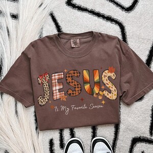 Op de afbeelding: Bruin t-shirt met het woord "JESUS" in een herfstthema. De letters zijn versierd met luipaardprint, ruitjes en herfstkleuren. De tekst "Is My Favorite Season" staat eronder.