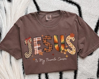 May include: Brown t-shirt featuring the word "JESUS" in a seasonal design. The letters are decorated with leopard print, plaid, and autumn colours. The text "Is My Favorite Season" is below the word JESUS.