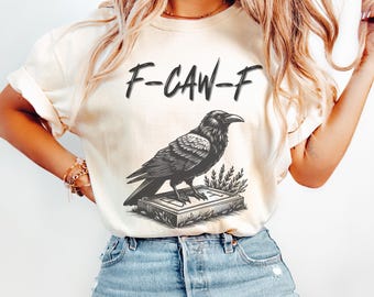 May include: Cream-colored t-shirt featuring a black illustration of a raven perched on a stone. The text "F-CAW-F" is above the raven. The raven is detailed with textured feathers and stands on a stone with a small plant.