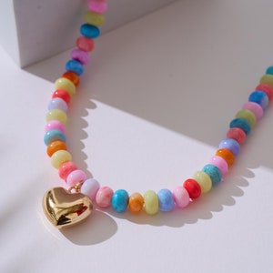 Puede incluir: Un collar de cuentas de colores con un colgante de corazón dorado. Las cuentas son de color rosa, azul, amarillo, verde y naranja.