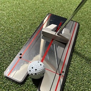 May include: A golf putting training aid featuring a silver putter, a white golf ball with black dimples, and a silver and black putting alignment tool with red lines. The aid is on a green surface.