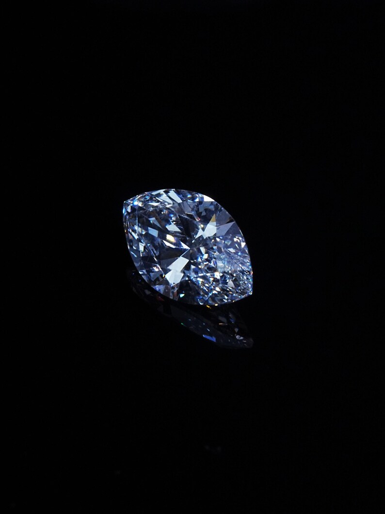 May include: A marquise-cut, light blue diamond against a black background. The diamond's facets reflect light, creating a sparkling effect. The gemstone is the focal point, with a dark, reflective surface beneath.