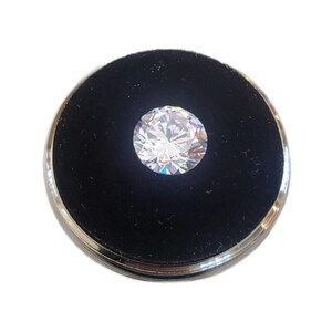 May include: A round, brilliant-cut diamond set in a black velvet display case with a silver-colored rim. The diamond sparkles with multiple facets, reflecting light. The case is circular and sits on a white background.