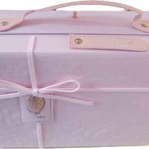 May include: A light pink, hexagonal-shaped storage box with a handle and a clasp. The box is adorned with a floral embossed pattern and tied with a pink ribbon. A small tag with a gold leaf charm hangs from the ribbon.