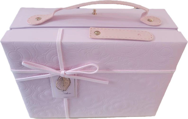 May include: A light pink, hexagonal-shaped storage box with a handle and a clasp. The box is adorned with a floral embossed pattern and tied with a pink ribbon. A small tag with a gold leaf charm hangs from the ribbon.