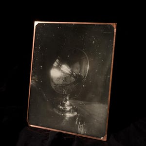 May include: A black and white photograph of a glass with a light bulb inside, mounted on a stand. The photograph is framed with a copper border. The background is black.