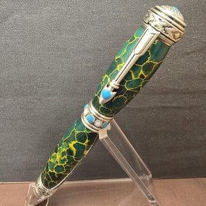 May include: A ballpoint pen featuring a dark green barrel with a yellow crackle pattern. The pen includes silver-toned metal accents, a clip shaped like an arrow, and small circular blue cabochon inlays along the center band and the top cap.