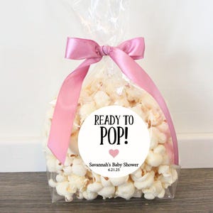 May include: A clear cellophane bag filled with popcorn, tied with a pink satin ribbon. A white circular sticker reads "READY TO POP!" with a pink heart and baby shower details.