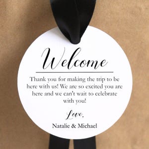 May include: A circular white gift tag with black text that reads Welcome, Thank you for making the trip to be here with us! We are so excited you are here and we can't wait to celebrate with you! Love, Natalie & Michael. The tag hangs from a black ribbon.