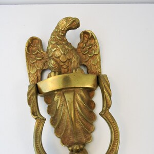 May include: A brass door knocker featuring an eagle perched above a banner. The knocker has a decorative leaf design and a curved handle. The eagle has detailed feathers and a proud pose. The overall design is ornate and vintage.