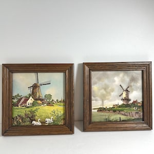 May include: Two framed paintings of Dutch windmills. The first painting shows a windmill in a rural setting with a small house and swans in the foreground. The second painting shows a windmill on a waterfront with a cloudy sky.
