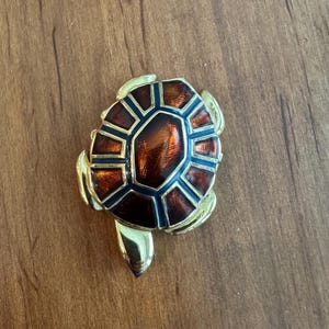 May include: A gold-toned turtle brooch with a detailed shell design. The shell features a geometric pattern in shades of brown and dark blue, with a central, glossy, amber-colored section. The brooch has a polished finish.