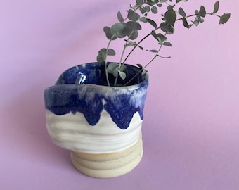 May include: A white ceramic vase with a blue glaze, shaped like a wave, with a sprig of eucalyptus leaves inside.