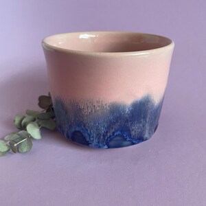 May include: A ceramic cup with a pink upper section and a dark blue, dripped glaze on the lower half. The cup has a smooth, rounded rim and a cylindrical shape. The background is a solid lilac colour.