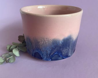 May include: A ceramic cup with a pink upper half and a dark blue, dripped glaze on the lower half. The cup has a smooth, rounded rim and a cylindrical shape. The background is a solid lavender color.