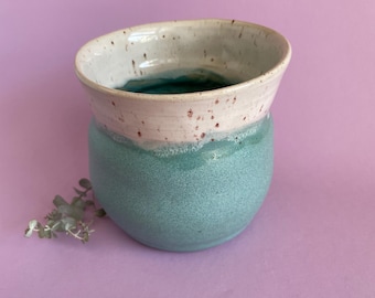 May include: A handmade ceramic vase with a rounded base and a wide, slightly flared rim. The lower portion is a speckled teal color, while the rim is a speckled off-white. The interior is a darker teal. A small sprig of greenery rests near the vase.