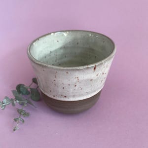 May include: A handcrafted ceramic cup with a white speckled upper section and a dark brown base. The cup has a wide opening and a slightly tapered form. The background is a solid light purple colour.