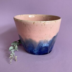 May include: A ceramic bowl with a pink upper section and a deep blue lower section. The bowl has a speckled texture and a slightly flared shape. The colours blend in a wave-like pattern. The bowl is a decorative item.
