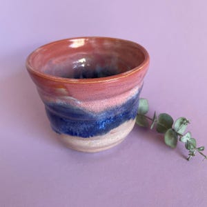 May include: A handcrafted ceramic cup with a layered design. The top rim is reddish-orange, transitioning to pink, then blue, and finally a cream base. A sprig of eucalyptus rests beside the cup on a purple surface.