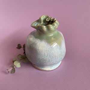 Peut inclure: Petit vase en céramique avec un corps arrondi et un dessus froncé. Le vase est émaillé dans des tons de vert pâle et de bleu, avec un effet moucheté. La base est de couleur beige clair. Une brindille de verdure repose à côté du vase.