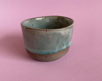 May include: A small, light blue ceramic cup with a brown rim. The cup has a speckled glaze and a slightly uneven surface.
