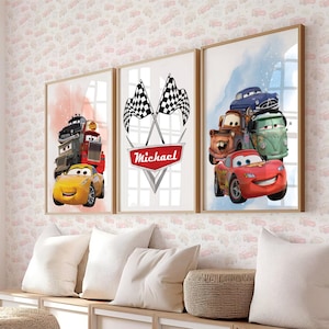 Op de afbeelding: Drie ingelijste prints met cartoonauto's. Eén print toont een gele auto, een rode vrachtwagen en een zwarte vrachtwagen tegen een roze en oranje achtergrond. Een andere print toont geblokte vlaggen en de naam "Michael". De derde print toont verschillende auto's tegen een blauwe achtergrond.