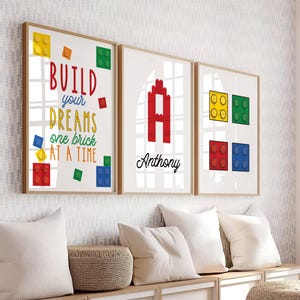 May include: Three framed art prints with a wooden frame. The prints feature colorful block designs and the text "BUILD your DREAMS one brick AT A TIME" and the name "Anthony". The prints are displayed above white pillows.