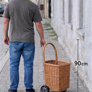 May include: A woven wicker shopping basket on wheels, approximately 90cm tall, with a curved handle. The basket is a natural light brown colour and is being pulled along a brick pavement.