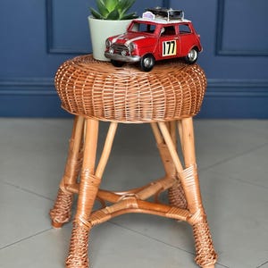 Peut inclure: Un petit tabouret en osier tressé avec un dessus rond et trois pieds. Une voiture jouet rouge avec le numéro 177 est posée dessus, ainsi qu'une petite plante en pot. Le tabouret est en osier et en bois de couleur naturelle.