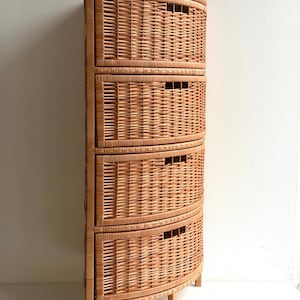 Pode incluir: Um armário alto e curvo de vime com quatro gavetas. O armário é feito de vime castanho claro tecido e tem uma forma semicircular. Cada gaveta tem uma abertura retangular para uma pega. O armário assenta em pernas curtas.